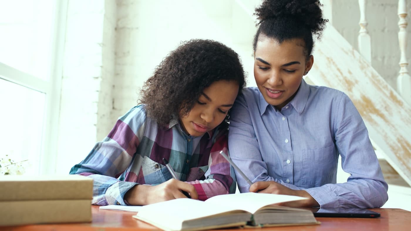 A parent helping a student with schoolwork at a table at home, both writing together