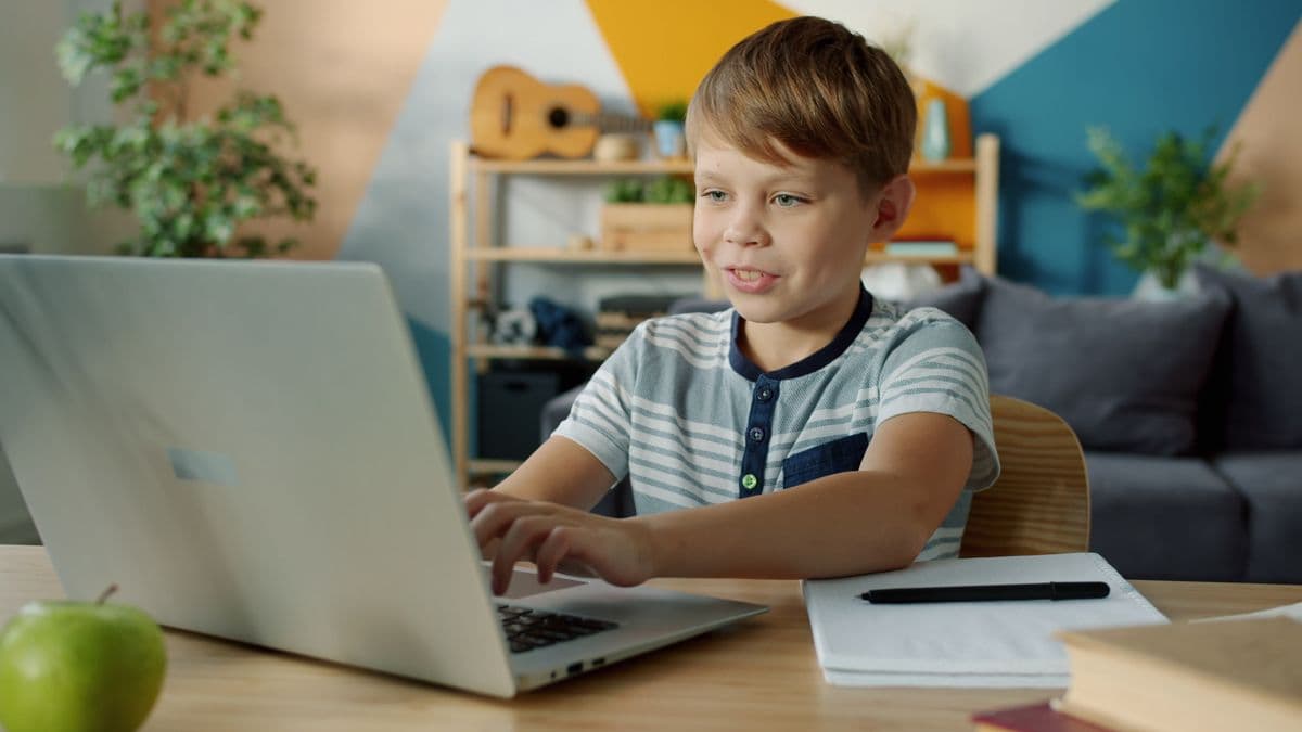 A happy young student typing on a laptop at a desk, a notebook and pen beside them